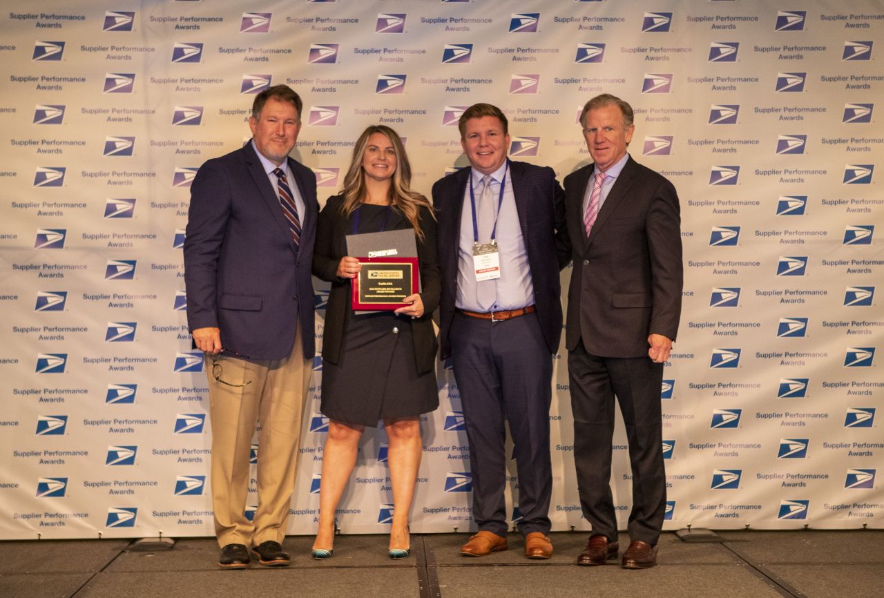 A man standing next to a woman holding an award with two men standing on her opposite side in front of a USPS backdrop. They are all smiling and dressed professionally.