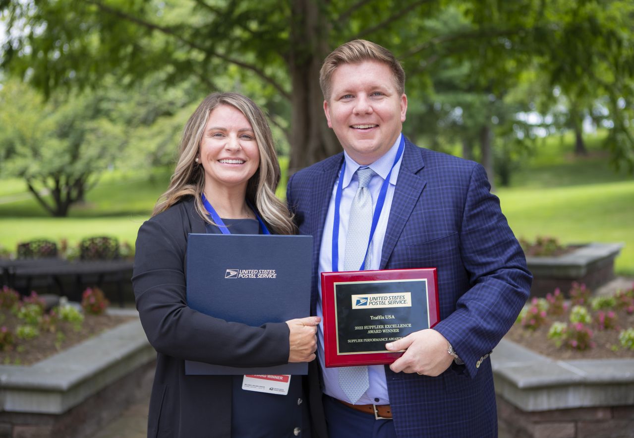 A professionally dressed woman and a man who work together standing in front of a lush green park, smiling, and holding an award.