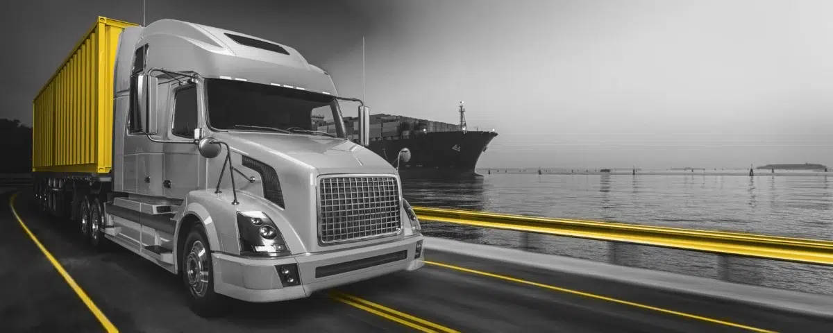 Black and white photo of a truck driving on a road. Shipping container colored yellow.