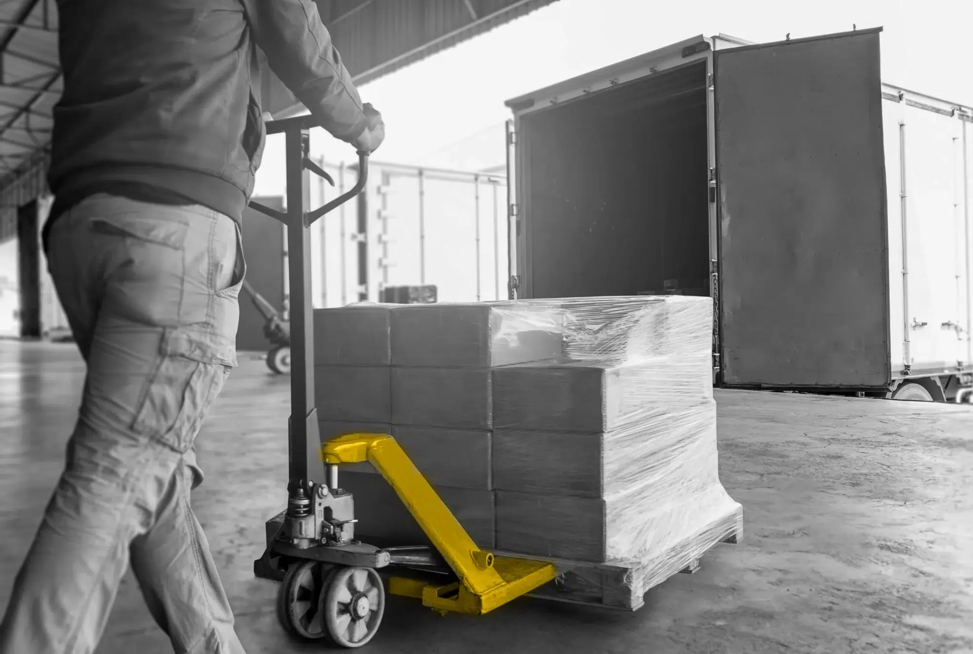 Black and white photo of pallets being loaded into a truck. Pallet loader colored yellow.